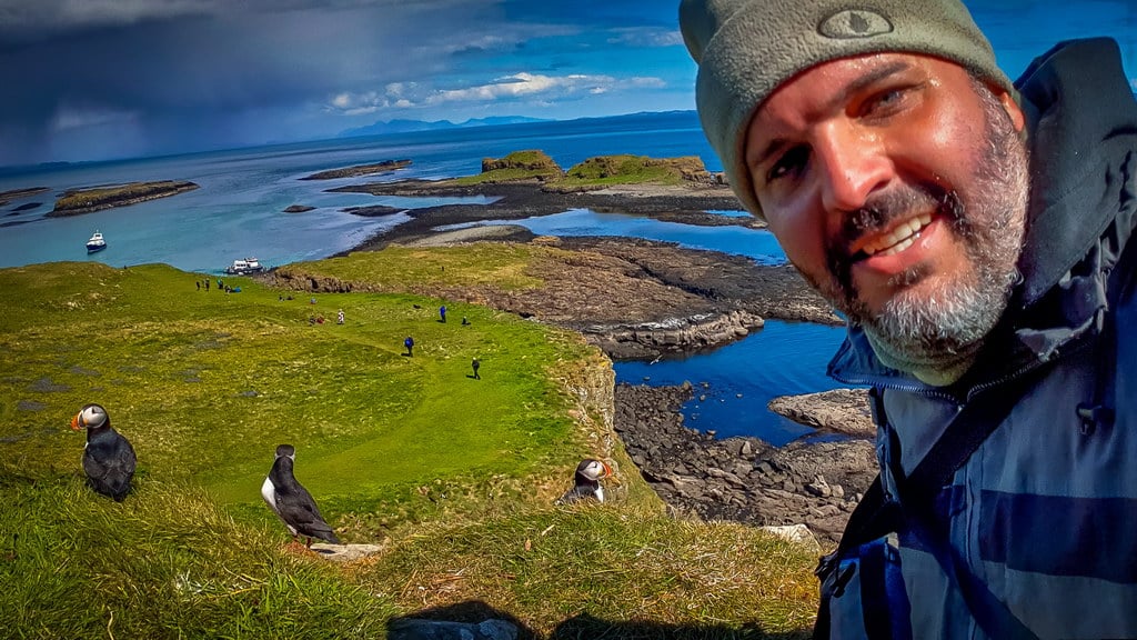 Observador de aves captura selfie na Escócia, com papagaios-do-mar ao fundo, em uma ilha remota."Finalidade da Imagem: A imagem ilustra um fotógrafo de natureza em viagem, enfatizando a beleza e o desafio de fotografar em locais remotos e exóticos como as ilhas da Escócia.